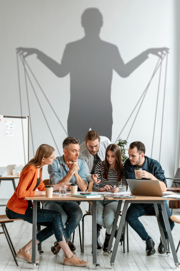 Nell'immagine un'ombra sinistra tira i fili di un gruppo di persone in un ufficio - Smart Marketing